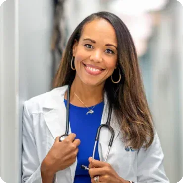 A woman wearing a white lab coat and a stethoscope around her neck stands indoors, smiling at the camera.