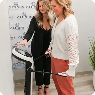 Two women stand by a body composition analyzer; one holds the handles while the other points at the device’s screen in a clinical setting.